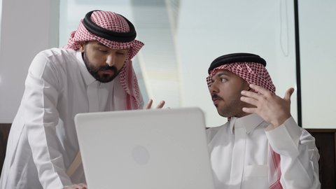 Discussion and explanation of statistical indicators and symbols, managing work using a laptop, a meeting of two Arab Saudi Gulf men wearing traditional attire and shemagh at the company headquarters, the Saudi work environment, unity and teamwork, achieving the company's goals, developing work and solving problems, Saudi office professions and jobs.