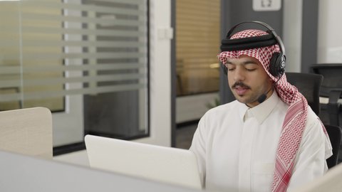 Responding to customer inquiries, working in the field of customer service and technical support, using headset technology, a photo of a Saudi Arabian Gulf man wearing traditional attire and a ghutrah, wearing a headset, sitting in an office in front of a laptop, using modern technology.