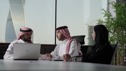 Conversations and discussions in the work environment, Arab Saudi Gulf employees wearing traditional attire and the ghutrah sitting in the meeting room inside the company headquarters, Saudi professions and jobs, a Saudi work team, following up and discussing the company's strategies and goals, completing required tasks via the laptop, integrating technology into work.