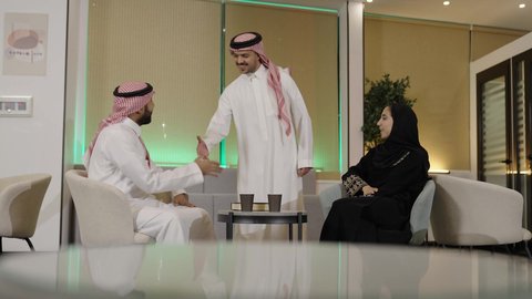 Collaboration in accomplishing tasks, two Arab Gulf men from Saudi Arabia wearing traditional thob and shemagh shaking hands with gestures of welcome and peace while greeting, accompanied by their female colleague, unity and collective interaction to achieve the company's goals and solve problems, office professions and jobs.