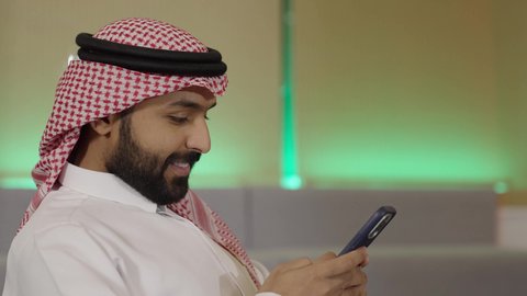 التواصل مع العملاء عن بعد, استخدام التقنيات الحديثة و المتطورة في العمل, مهن و وظائف في السعودية, تصوير مقرب من الجانب لموظف عربي خليجي سعودي يرتدي الثوب التقليدي و الشماغ يجلس داخل المكتب و يمسك الهاتف المحمول بيده, أعمال إدارية مكتبية