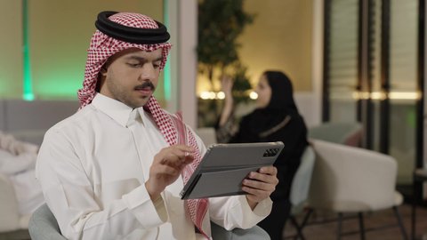 تصوير مقرب  لموظف عربي خليجي سعودي يرتدي الثوب التقليدي والشماغ يحمل بيده جهاز لوحي في المكتب،  ايماءات الاندماج و التركيز، استعمال التكنولوجيا في العمل عن بعد، استخدام الأجهزة الحديثة في انجاز الأعمال، تصفح مواقع التواصل الاجتماعي عن طريق الانترنت