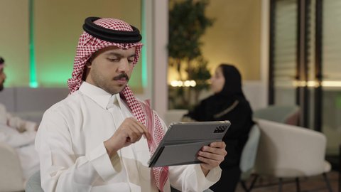 Gestures of integration and focus, using technology for remote work, utilizing modern devices to accomplish tasks, browsing social media sites via the internet, a close-up shot of a Saudi Gulf Arab employee wearing traditional attire and a ghutrah holding a tablet in the office.