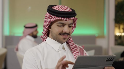 استخدام الأجهزة الحديثة في انجاز الأعمال، تصفح مواقع التواصل الاجتماعي عن طريق الانترنت، استعمال التكنولوجيا في العمل عن بعد،تصوير مقرب  لموظف عربي خليجي سعودي يرتدي الثوب التقليدي والشماغ يحمل بيده جهاز لوحي في المكتب، ايماءات الاندماج و التركيز