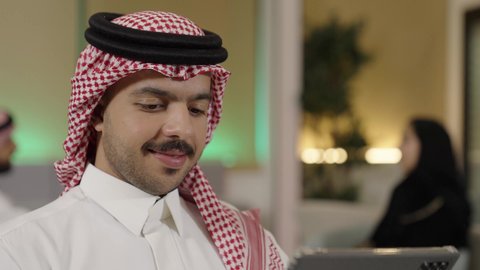 تصفح مواقع التواصل الاجتماعي عن طريق الانترنت، استخدام الأجهزة الحديثة في انجاز الأعمال، استعمال التكنولوجيا في العمل عن بعد،تصوير مقرب  لموظف عربي خليجي سعودي يرتدي الثوب التقليدي والشماغ يحمل بيده جهاز لوحي في المكتب، ايماءات الاندماج و التركيز