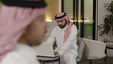 استخدام الأجهزة التقنية الحديثة في مكاتب الشركات، تصوير مقرب لوجه رجل عربي خليجي سعودي يرتدي الشماغ يتحدث الى شخص ما ، شاب يعمل على جهاز الحاسوب في مقر العمل، انجاز مهام العمل اونلاين