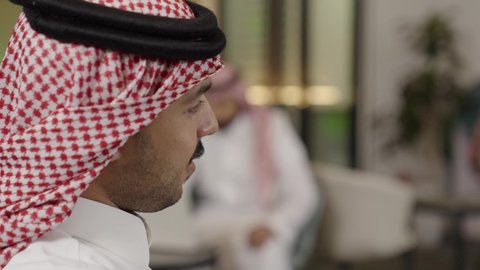 تصوير مقرب لوجه رجل عربي خليجي سعودي يرتدي الشماغ يتحدث الى شخص ما ، شاب يعمل على جهاز الحاسوب في مقر العمل، استخدام الأجهزة التقنية الحديثة في مكاتب الشركات، انجاز مهام العمل اونلاين