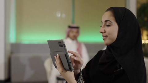 النقر على شاشة التابلت، استخدام الأجهزة التقنية الحديثة، مفهوم إنجاز المشاريع والمهام العمل عن بعد، تصفح مواقع التواصل الاجتماعي، التواصل مع الأهل والأصدقاء عبر الايباد، تصوير مقرب من الجانب لإمرأة عربية خليجية سعودية محجبة ترتدي العباءة السوداء تقوم باستخدام الجهاز اللوحي