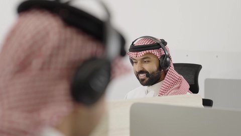 Video calls during a work meeting, a Saudi Gulf Arab man wearing traditional attire and a headscarf, wearing headphones, sitting in an office working in customer service, responding to inquiries and questions, using modern and advanced technologies.