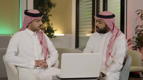 تطوير ودراسة المشاريع الجديدة في شركات المملكة العربية، استخدام جهاز تقني حديث، جمع البيانات وحل المشكلات، اجتماع ومناقشة عمل في مقر الشركة، موظف عربي خليجي سعودي  الثوب الابيض و الشماغ يجلس امام الحاسب المحمول داخل المكتب برفقة زميله