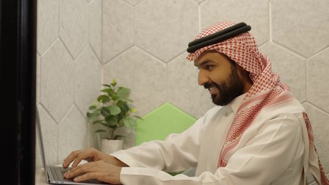 استخدام الأجهزة التقنية الحديثة في مكاتب الشركات، إدارة الأعمال المكتبية وتنظيمها، النقر والتفاعل مع لوحة المفاتيح،  تصوير مقرب من الجانب  لموظف  عربي خليجي سعودي يرتدي الثوب القليدي و الشماغ يقوم بإنجاز مهام العمل المطلوبة عبر الحاسب المحمول