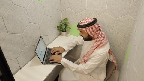 النقر والتفاعل مع لوحة المفاتيح، إدارة الأعمال المكتبية وتنظيمها،  تصويرمن الجانب  لموظف  عربي خليجي سعودي يرتدي الثوب القليدي و الشماغ يقوم بإنجاز مهام العمل المطلوبة عبر الحاسب المحمول، استخدام الأجهزة التقنية الحديثة في مكاتب الشركات