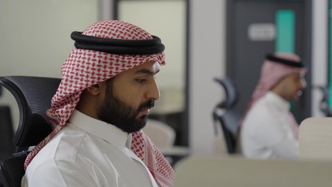 بيئة العمل المريحة في شركات السعودية ، إدارة الأعمال المكتبية وتنظيمها، تصويرمقرب من الجانب لرجل عربي خليجي سعودي يرتدي الزي التقليدي و الشماغ يعمل بايماءات الاندماج و التركيز، وظائف ومهن إدارية سعودية