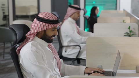 استخدام التكنولوجيا في الحياة العملية، تصوير من الجانب لموظفون عربيون خليجيون سعوديون يرتدون الزي التقليدي و الشماغ يعملون على الحاسب المحمول في مقر الشركة، بيئة العمل المريحة في شركات السعودية ، إدارة الأعمال المكتبية وتنظيمها