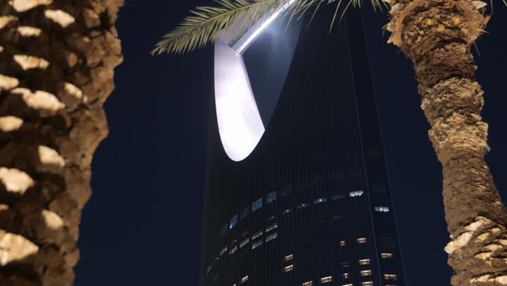 The advanced architectural engineering art of the illuminated Kingdom Tower building in the capital of Saudi Arabia, the development of modern engineering facilities in the Kingdom, famous tourist places and landmarks to attract visitors from all over the world, skyscrapers at night in the city of Riyadh, green palm trees.