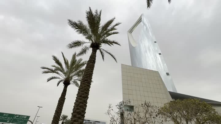 Famous tourist places and landmarks to attract tourists from all over the world, the development of architectural engineering art in a modern style, a side view of the famous Kingdom Tower in Riyadh, towers and skyscrapers, green palm trees, road signs to facilitate movement in the city.