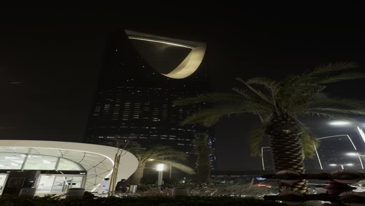 Night photography of a famous landmark in the capital, green palm trees in the area of the famous Kingdom Tower in Riyadh, well-known tourist spots and attractions to attract visitors from all over the world, the evolution of architectural engineering art in a modern style, towers and skyscrapers.