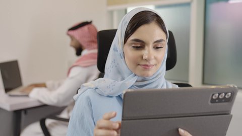 Integrating modern and advanced technologies into practical life, collaboration among colleagues, office professions and jobs, a close-up shot of a Saudi Gulf Arab woman sitting in the company headquarters working on a tablet, in the background a Saudi man completing daily tasks on a laptop.