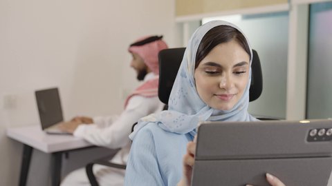 استخدام التقنيات الحديثة والمتطورة في الحياة العملية, التشارك بالعمل بين الزملاء, مهن ووظائف مكتبية, تصوير مقرب لإمراة عربية خليجية سعودية تجلس في مقر الشركة تعمل على جهاز اللوحي المحمول, خلفية رجل سعودي يقوم بانجاز المهام اليومية على الحاسب المحمول