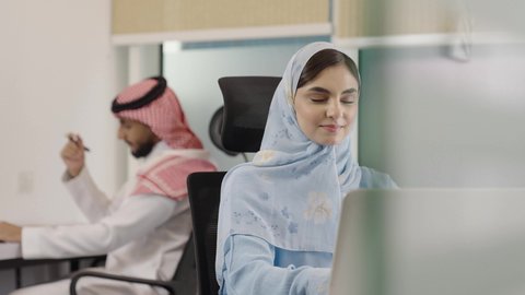 Equality between men and women, using modern technologies in professional life, sharing work among colleagues, office professions and jobs, a close-up shot of a Saudi Gulf Arab woman sitting in the company headquarters working on a laptop, in the background a Saudi man is recording and taking notes on a paper.