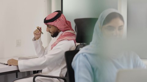العمل الجماعي وروح الفريق الواحد، استخدام التقنيات الحديثة في الحياة العملية, التعاون بالعمل بين الزملاء, مهن ووظائف مكتبية, تصوير مقرب لإمراة عربية خليجية سعودية تجلس في مقر الشركة تعمل على جهاز الحاسب المحمول, خلفية رجل سعودي يقوم بتسجيل وتدوين الملاحظات