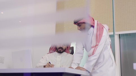 تدوين الملاحظات على الورق، الوحدة والتفاعل الجماعي لتحقيق أهداف الشركة وحل المشكلات، وظائف مكتبية، الاهتمام بالمظهر الخارجي ومفهوم الاناقة، استخدام جهاز تقني حديث ومتطور، رجلين عربيين خليجيين سعوديين يرتديان الشماغ والثوب التقليدي يتناقشون في مكتب أعمال