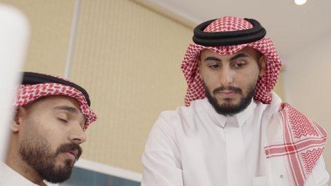 Dialogue in task completion, unity and collective interaction to achieve company goals and solve problems, office functions, attention to external appearance and the concept of elegance, using modern and advanced technical devices, a close-up shot of two Arab Gulf Saudi men wearing the ghutrah and traditional thobe discussing in a business office.