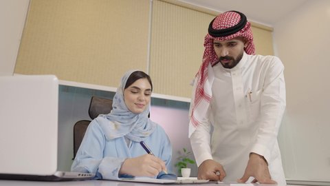 تسجيل ملاحظات لتحقيق الاستراتيجيات والغايات، تبادل الآراء بين الزملاء، إدارة شؤون الأعمال عبر الاجهزة الحديثة ، دمج التقنيات المتطوره فى الحياه اليوميه، رجل عربي خليجي سعودي يقوم بالشرح عن مشروع ،إمرأة سعودية ترتدي العباءة والحجاب تستمع إلى ملاحظات مهمة