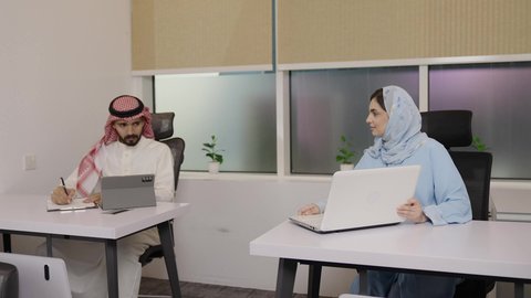 المناقشة بين الزملاء بالمهام الفردية، استخدام التقنيات الحديثة في الحياة العملية, التعاون بالعمل بين الزملاء, مهن ووظائف مكتبية, إمراة عربية خليجية سعودية تجلس في مقر الشركة تعمل على جهاز الحاسب المحمول, رجل سعودي يقوم بتسجيل وتدوين الملاحظات على ورقة