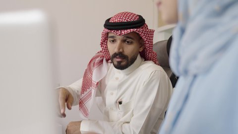 الاستشارة بين الزملاء عن المهام الفردية، استخدام التقنيات الحديثة في الحياة العملية, التعاون بالعمل بين الزملاء, مهن ووظائف مكتبية, إمراة عربية خليجية سعودية تجلس في مقر الشركة تعمل على جهاز الحاسب المحمول, رجل سعودي يقوم بمساعدة زميلته وتقديم النصائح