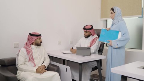 كتابة وتدوين البيانات والنتائج، ادارة وانجاز المهام باستخدام جهاز تقني حديث ومتطور, العمل على الجهاز اللوحي، مهنة ووظيفة مكتبية سعودية، رجلين عربيين خليجيين سعوديين يرتديان الشماغ والثوب التقليدي وتقف بجانبهما السكرتيرة تقدم الملفات المطلوبة