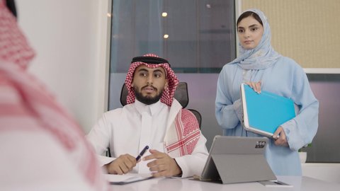 Managing and completing tasks using a modern and advanced technical device, writing and recording data and results, working on a tablet, a profession and office job in Saudi Arabia, a close-up shot of two Arab Gulf Saudi men wearing the traditional ghutrah and thobe, with a secretary standing next to them presenting the required files.