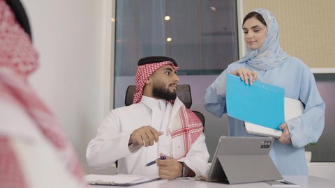 العمل على الجهاز اللوحي، مهنة ووظيفة مكتبية سعودية، كتابة وتدوين البيانات والنتائج، ادارة وانجاز المهام باستخدام جهاز تقني حديث ومتطور, تصوير مقرب لرجلين عربيين خليجيين سعوديين يرتديان الشماغ والثوب التقليدي وتقف بجانبهما السكرتيرة تقدم الملفات المطلوبة