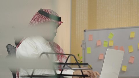 انجاز المهام المكتبية عبر التقنيات الالكترونية الحديثة والمتطورة, ادارة الأعمال عبر الاونلاين, مهن ووظائف مكتبية سعودية, بيئة العمل المريحة في الشركات الخليجية الراقية, رجل عربي خليجي سعودي يرتدي الثوب التقليدي والشماغ يعمل على الحاسب المحمول في المكتب