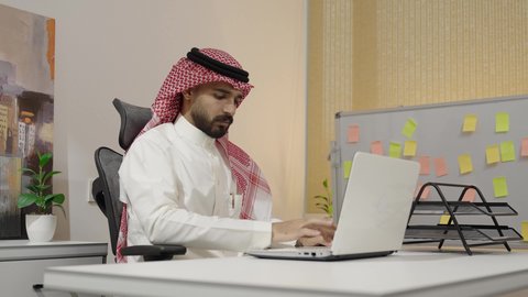 ادارة الأعمال عبر الاونلاين, انهاء المهام المكتبية عبر التقنيات الالكترونية الحديثة والمتطورة, مهن ووظائف مكتبية سعودية, بيئة العمل المريحة في الشركات الخليجية الراقية, رجل عربي خليجي سعودي يرتدي الثوب التقليدي والشماغ يعمل على الحاسب المحمول في المكتب