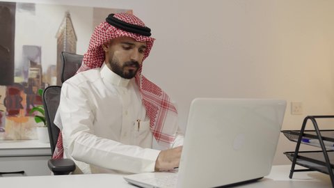 Monitoring work online, completing office tasks through modern and advanced electronic technologies, Saudi office professions and jobs, the comfortable work environment in upscale Gulf companies, an Arab Gulf Saudi man wearing traditional attire and a shemagh working on a laptop in the office.