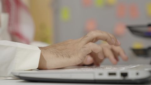 Fast typing skills on the keyboard, managing work online, completing office tasks using modern and advanced electronic technologies, an Arab Gulf Saudi man wearing traditional attire and a shemagh working on a laptop in the office, Saudi office jobs and professions.