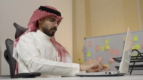 مهن ووظائف مكتبية سعودية, متابعة الأعمال عبر الاونلاين, انهاء المهام المكتبية عبر التقنيات الالكترونية الحديثة والمتطورة,  بيئة العمل المريحة في الشركات الخليجية الراقية, رجل عربي خليجي سعودي يرتدي الثوب التقليدي والشماغ يعمل على الحاسب المحمول في المكتب