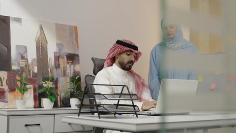 حوارات ومناقشات لتحقيق الاستراتيجيات والغايات، تبادل الآراء بين الزملاء، إدارة شؤون الأعمال عبر الاجهزة الحديثة ، دمج التقنيات المتطوره فى الحياه اليوميه، رجل عربي خليجي سعودي يقوم بالشرح عن مشروع ،إمرأة سعودية ترتدي العباءة والحجاب تستمع إلى ملاحظات مهمة