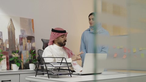مناقشات وحوارات لتحقيق الاستراتيجيات والغايات، تبادل الآراء بين الزملاء، إدارة شؤون الأعمال عبر الاجهزة الحديثة ، دمج التقنيات المتطوره فى الحياه اليوميه، رجل عربي خليجي سعودي يقوم بالشرح عن مشروع ،إمرأة سعودية ترتدي العباءة والحجاب تستمع إلى ملاحظات مهمة