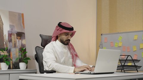 The comfortable work environment in upscale Gulf companies, monitoring business online, completing office tasks through modern and advanced electronic technologies, an Arab Gulf Saudi man wearing traditional attire and a shemagh working on a laptop in the office, Saudi office jobs and professions.