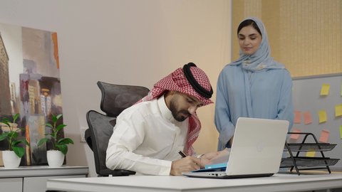 The comfortable work environment in Saudi companies, collaboration and exchange of opinions among colleagues, managing and organizing business affairs through modern devices, integrating advanced technologies into daily life, an Arab Gulf Saudi woman wearing an abaya and hijab carrying a signed work file, a Saudi man signing a file.
