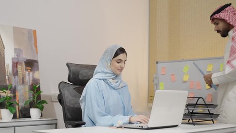 Collaboration and exchange of opinions among colleagues, managing and organizing business affairs through modern devices, integrating advanced technologies into daily life, an Arab Gulf Saudi woman wearing an abaya and hijab signing a file for her colleague at work, a Saudi man presenting a work file for review.
