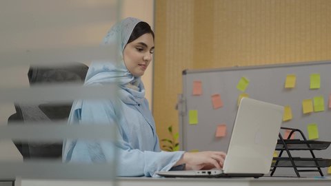 تسجيل الملاحظات على الحاسب المحمول، دمج التكنولوجيا والتقنيات الحديثة مع العمل، وظيفة مكتبية في شركات الخليج العربي، مفهوم الاناقة والاهتمام بالمظهر الخارجي،  امراة عربية خليجية سعودية محجبة ترتدي العباءة تجلس خلف الطاولة تعمل لانجاز المهام في المكتب