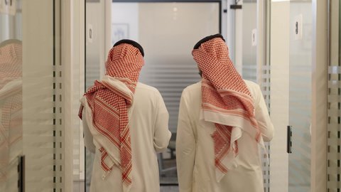 وظائف مكتبية، الاهتمام بالمظهر الخارجي ومفهوم الاناقة، استخدام جهاز تقني حديث ومتطور، تصوير من الخلف رجلين عربيين خليجيين سعوديين يرتديان الشماغ والثوب التقليدي يسيرون في ممر المكتب ويتناقشون في الأعمال، الوحدة والتفاعل الجماعي لتحقيق أهداف الشركة وحل المشكلات