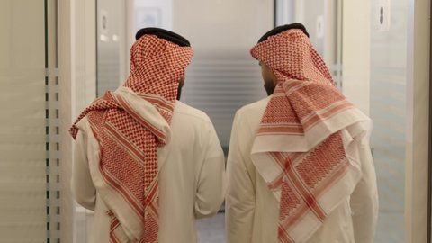 الوحدة والتفاعل الجماعي لتحقيق أهداف الشركة وحل المشكلات، وظائف مكتبية، الاهتمام بالمظهر الخارجي ومفهوم الاناقة، استخدام جهاز تقني حديث ومتطور، تصوير من الخلف رجلين عربيين خليجيين سعوديين يرتديان الشماغ والثوب التقليدي يسيرون في ممر المكتب ويتناقشون في الأعمال