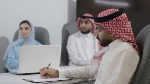 Taking important notes in a meeting, the comfortable work environment in Saudi companies, the use of modern technological devices in offices, discussions to achieve strategies and goals, a work meeting of Arab Gulf Saudi employees in the meeting room, understanding and cooperation among colleagues to achieve the company's objectives.