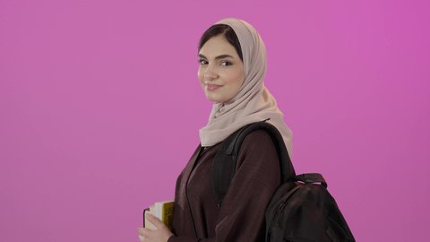 ايماءات الثقة بالنفس، التعليم في المملكة العربية السعودية, بورتريه لطالبة عربية سعودية خليجية ترتدي العباءة و حقيبة الظهرتحمل بيديها مجموعه من الكتب، النظر الى الكاميرا بايماءات السعادة و السرور, خلفية ملونه