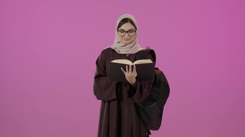 ايماءات الاندماج و التركيز، قراءة الكتب الجامعية، بورتريه طالبة جامعية عربية خليجية سعودية ترتدي العباءة و نظارة طبية تضع الحقيبة على ظهرها تحمل بيدها كتاب جامعي، مراجعة الدروس اليومية، التعليم الجامعي في السعودية ، خلفية ملونه