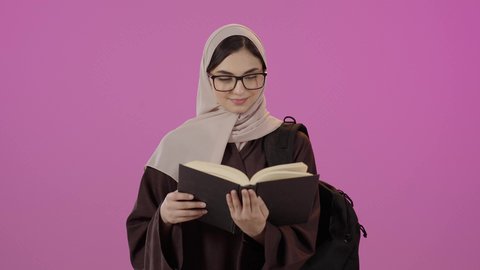 قراءة الكتب الجامعية، بورتريه طالبة جامعية عربية خليجية سعودية ترتدي العباءة و نظارة طبية تضع الحقيبة على ظهرها تحمل بيدها كتاب جامعي، مراجعة الدروس اليومية، التعليم الجامعي في السعودية ، خلفية ملونه